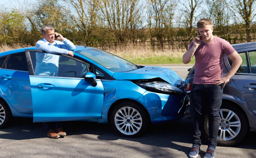 Two men on their phones after a rear-end car accident on a rural road in Texas, with visible damage to the front of a blue car and the rear of a gray car.