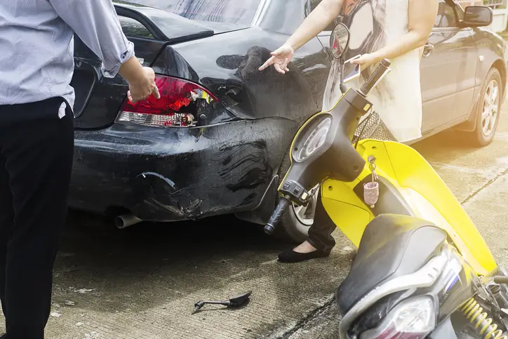 Two people pointing at a damaged car and a fallen yellow scooter after a collision on a city street